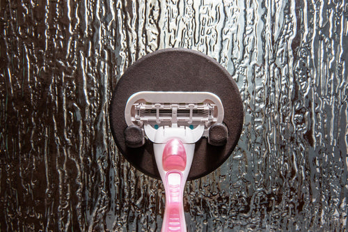Pink razor held by a round magnetic razor hook on textured shower glass