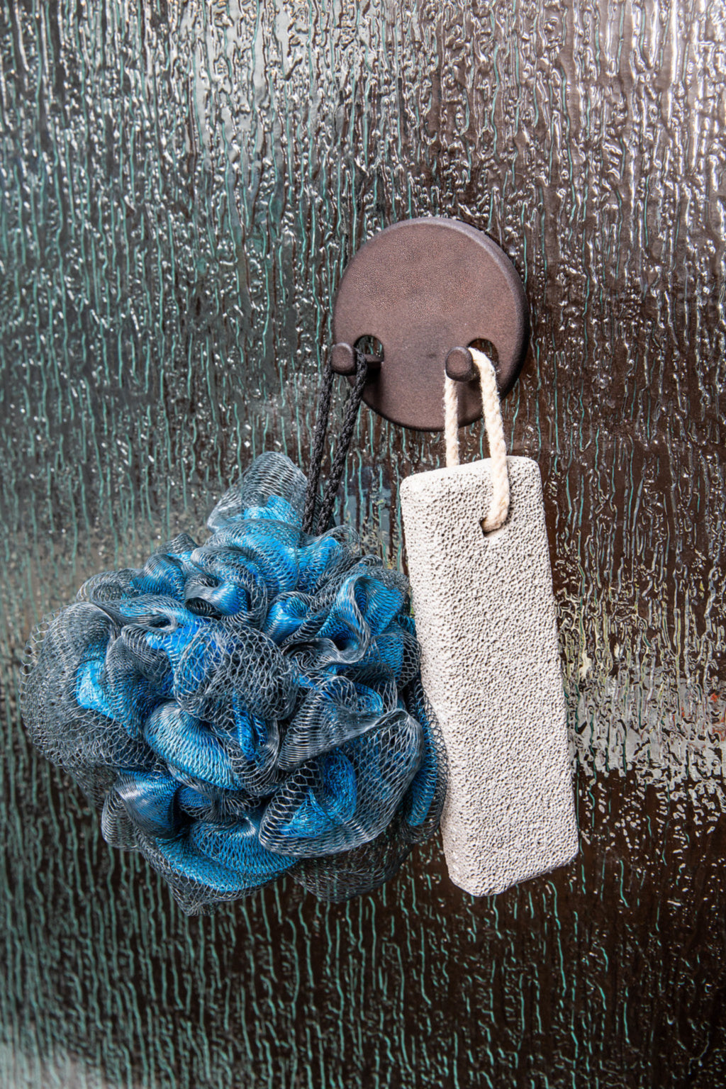 Bath pouf and pumice stone hanging from a magnetic hook on a glass shower door, used as a loofah hanger