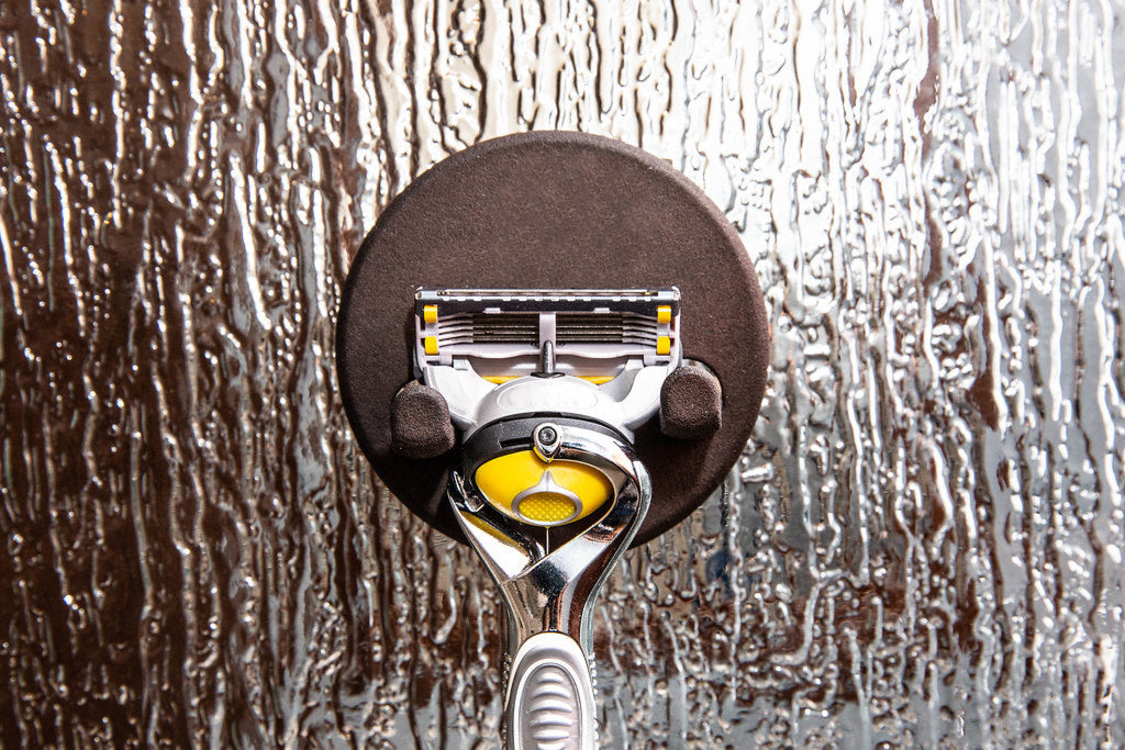 Silver razor with a yellow head held by a magnetic hook on frosted shower glass
